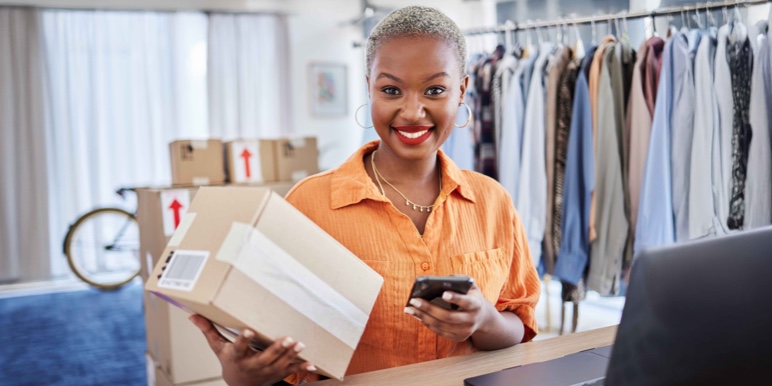 Smiling female business owner in an orange shirt holding a package and using a smartphone in a boutique clothing store. Shelves with boxes and racks of clothes suggest a thriving retail business, secured with business insurance.