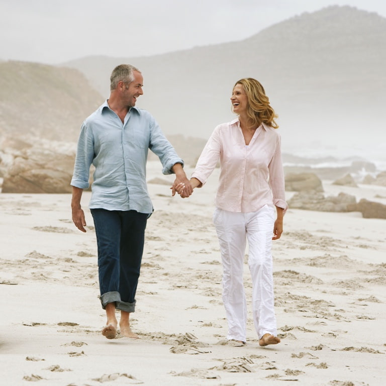 A financially secure, middle-aged couple holding hands and walking barefoot along the beach in overcast weather.