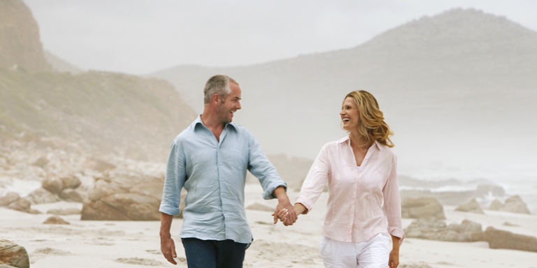 A financially secure, middle-aged couple holding hands and walking barefoot along the beach in overcast weather.