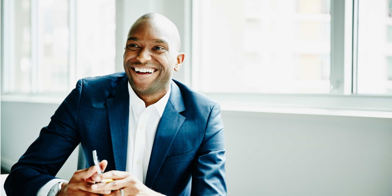 A laughing young African male professional at a desk, in a suit with a mobile phone.